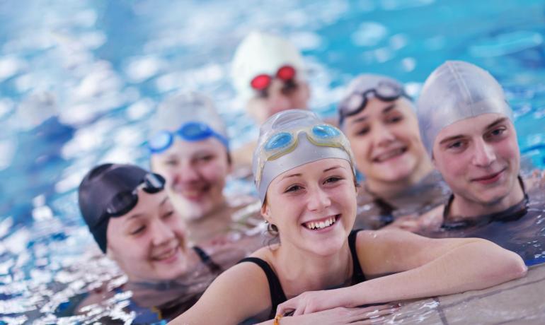 Gruppo di nuotatori sorridenti in piscina con cuffie e occhialini.