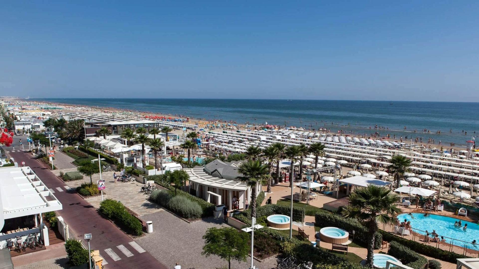 Spiaggia affollata con ombrelloni, piscina e pista ciclabile lungo la costa.