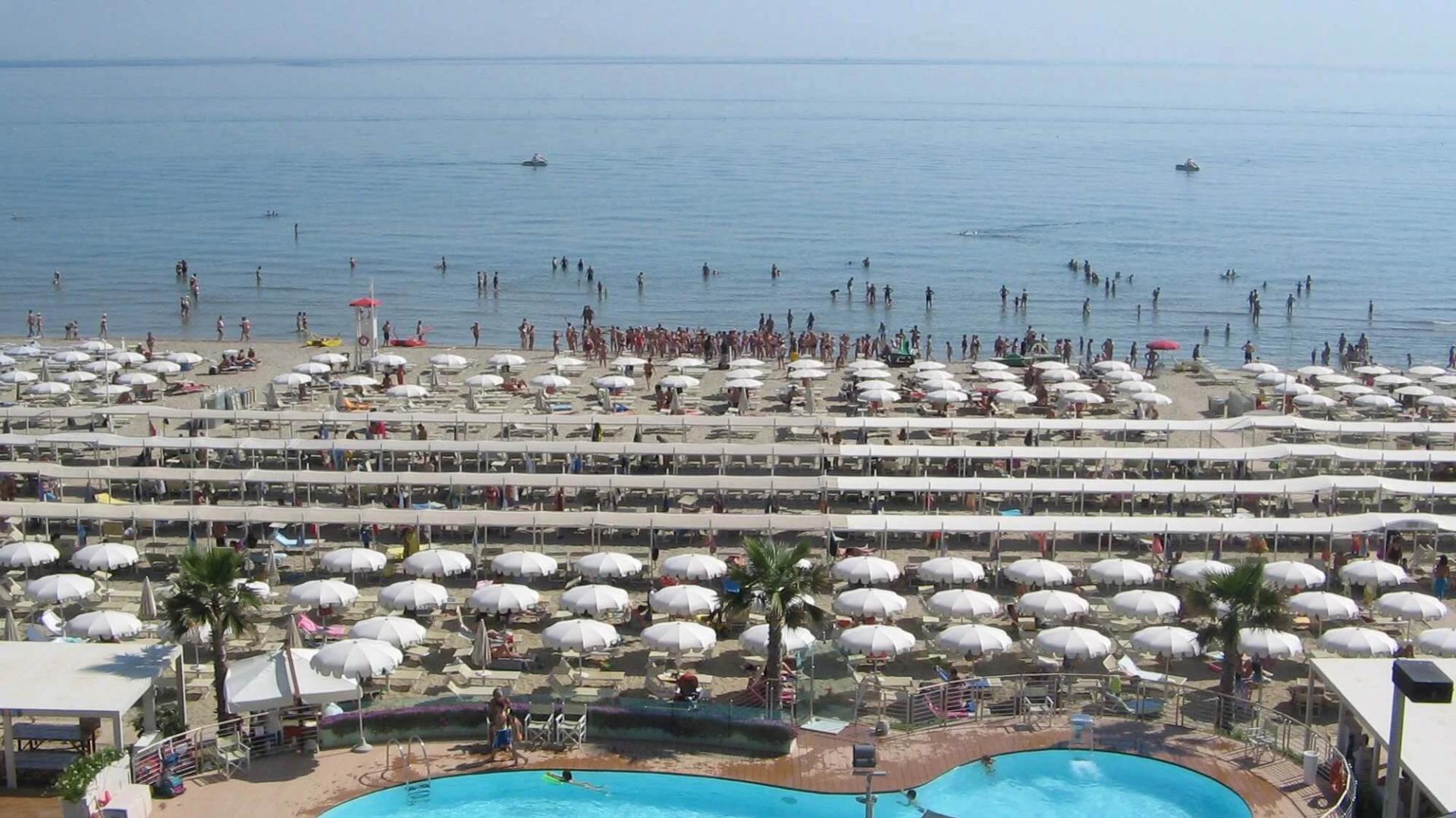 Spiaggia affollata con ombrelloni bianchi e piscina vicino al mare.